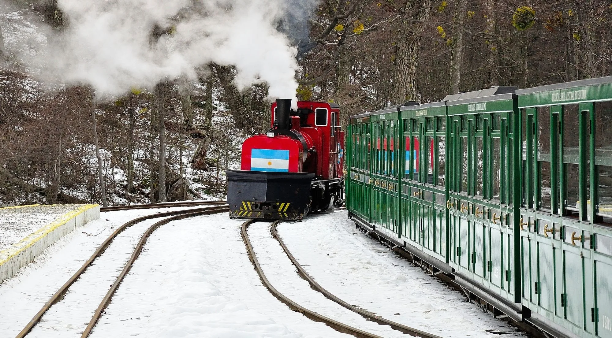 Red steam locomotive with an Argentine flag leading green carriages through snow