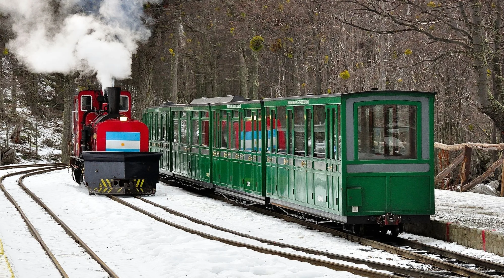 The End of the World Train's red steam locomotive and green carriages on a snow-covered track in winter